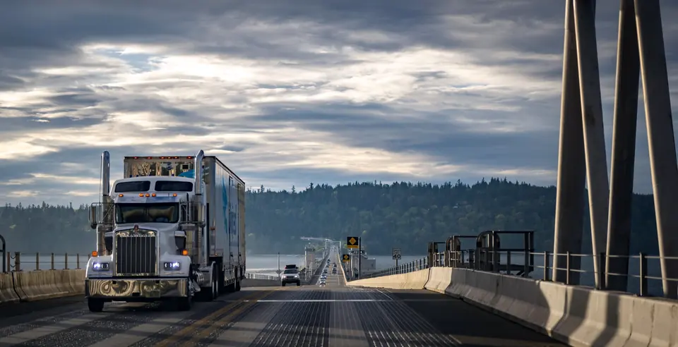 White semi truck hauling a refrigerated trailer across a highway bridge under a cloudy sky